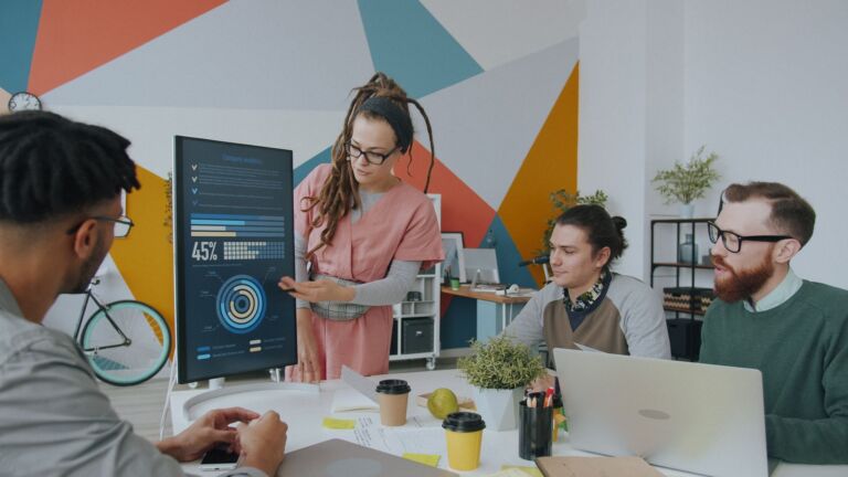 Dans un environnement de bureau dynamique, une équipe aux profils variés discute des analyses affichées sur un écran, favorisant ainsi la collaboration.