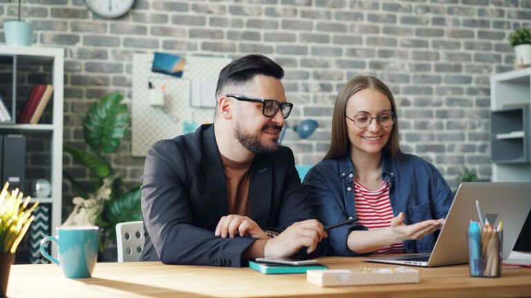 Un couple professionnel souriant et collaborant sur un ordinateur portable à une table de bureau moderne.