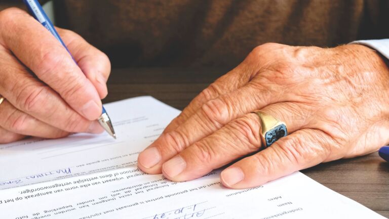 Gros plan d'un senior signant un document légal, mettant l'accent sur la main et l'alliance en or.